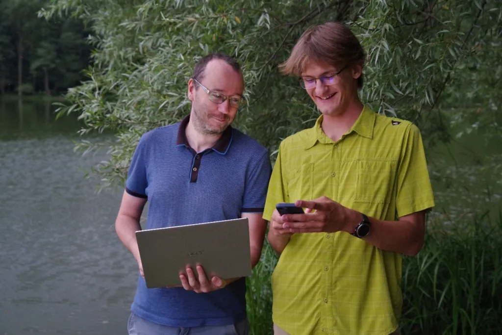 Dva muži u vody, jeden drží notebook a druhý se usmívá na telefon